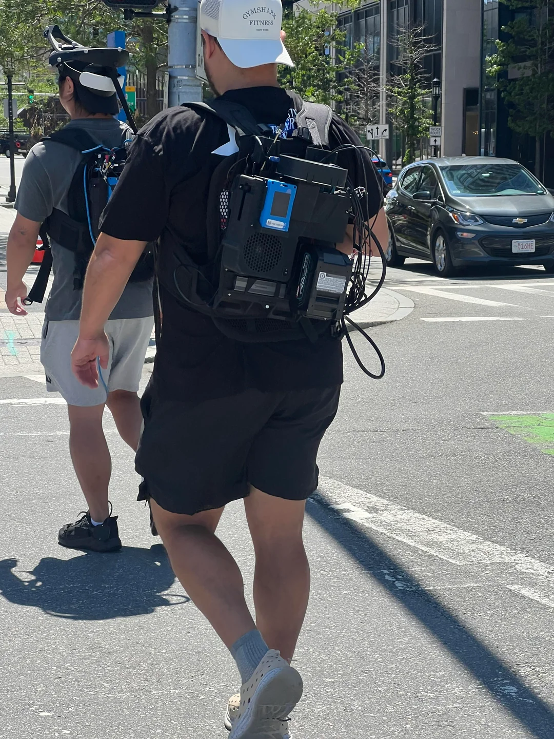 Man wearing a black backpack rig with visible electronics, fans and cables running to a head-mounted camera array, crossing a city street