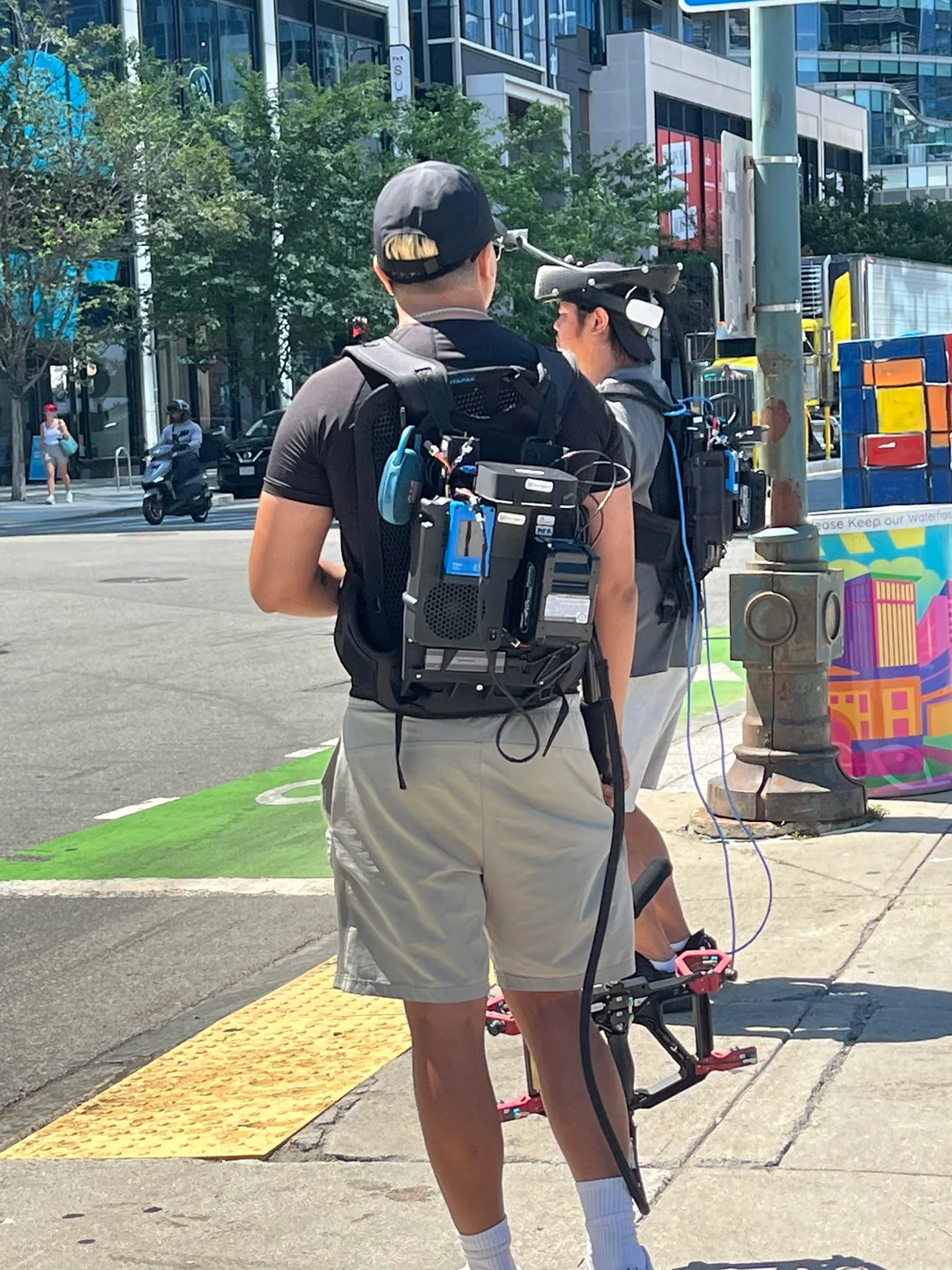 Two men wearing backpack sensor rigs with head-mounted camera arrays, one holding a handheld gimbal, on a city street corner